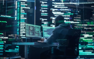 A man is sitting at a desk with computers and he's working. All around the room, there seems to be holographic-like projections of the work on his screen. It can be assumed to be code and IT specialty work.