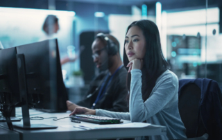 Cyber breach containment services. The image shows a woman seated at a desk, closely monitoring multiple large computer screens. She appears focused, with one hand resting on her chin and the other near the keyboard. Behind her, there is a man also working at a computer, wearing headphones. The room has a high-tech, professional atmosphere, suggesting a cybersecurity or IT environment. The lighting is cool and dim, and there are other people visible in the background, though blurred. The image conveys a sense of concentration and teamwork in a modern, technology-driven setting.