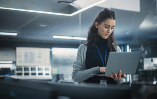 Effective Cyber Breach Containment Services. A Woman with brown hair stands with a silver laptop in hand and she appears to be typing something on it. She is in a gray office space.