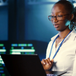 Data recovery post-ransomware. The image shows a woman standing in what appears to be a high-tech or server room environment, as there are monitors and screens with data displays in the background. She is focused on a laptop in front of her, appearing to work or analyze information. The woman is wearing glasses, a light-colored blouse, and a lanyard around her neck, suggesting a professional setting. The background has a blue tone, adding to the high-tech atmosphere, possibly indicating that she works in IT, cybersecurity, or data analysis.