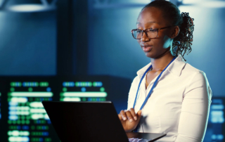 Data recovery post-ransomware. The image shows a woman standing in what appears to be a high-tech or server room environment, as there are monitors and screens with data displays in the background. She is focused on a laptop in front of her, appearing to work or analyze information. The woman is wearing glasses, a light-colored blouse, and a lanyard around her neck, suggesting a professional setting. The background has a blue tone, adding to the high-tech atmosphere, possibly indicating that she works in IT, cybersecurity, or data analysis.