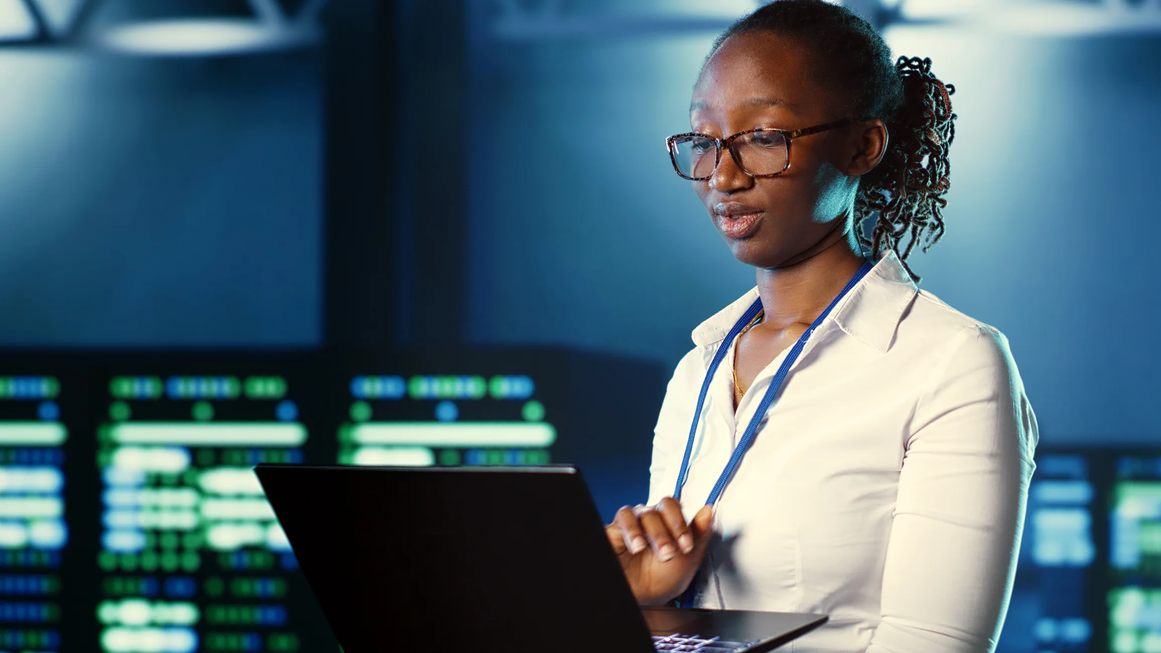 Data recovery post-ransomware. The image shows a woman standing in what appears to be a high-tech or server room environment, as there are monitors and screens with data displays in the background. She is focused on a laptop in front of her, appearing to work or analyze information. The woman is wearing glasses, a light-colored blouse, and a lanyard around her neck, suggesting a professional setting. The background has a blue tone, adding to the high-tech atmosphere, possibly indicating that she works in IT, cybersecurity, or data analysis.