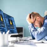 Incident Response Readiness. The image shows a man sitting at a desk, visibly stressed, with his hands on his head. He is staring at two computer monitors displaying warning messages that read "SYSTEM HACKED," suggesting that his computer system has been compromised. The room appears to be an office setting, with a light background and organized desk supplies. The man seems overwhelmed, likely due to the cyberattack that has disrupted his work.