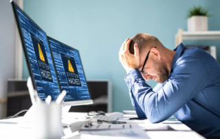 Incident Response Readiness. The image shows a man sitting at a desk, visibly stressed, with his hands on his head. He is staring at two computer monitors displaying warning messages that read "SYSTEM HACKED," suggesting that his computer system has been compromised. The room appears to be an office setting, with a light background and organized desk supplies. The man seems overwhelmed, likely due to the cyberattack that has disrupted his work.