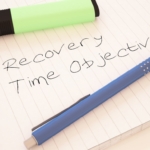 Recovery Time Objective. The image shows a lined notebook page with the handwritten phrase "Recovery Time Objective." A blue pen and a green highlighter are placed on the notebook, adding a casual yet purposeful feel to the setting. The phrase "Recovery Time Objective" emphasizes the importance of setting goals for system recovery after disruptions, a concept often used in business continuity and disaster recovery planning. The items suggest a planning or brainstorming session focused on determining acceptable downtime limits.