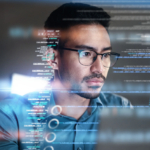 Ransomware recovery best practices. The image shows a man wearing glasses, intensely focused on a computer screen. He appears to be a programmer or someone working with code, as lines of code and data seem to float in front of him as a digital overlay. The man has short, dark hair, facial hair, and a thoughtful expression as he examines the information. The scene is futuristic, with a soft glow reflecting off the code, evoking a sense of technology and problem-solving, possibly in a cybersecurity or programming environment.