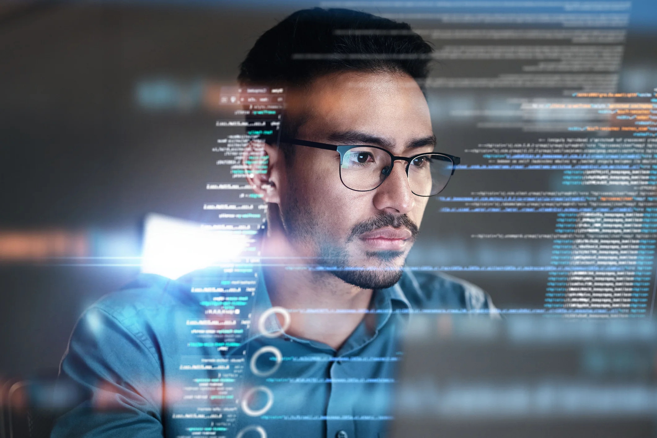 Ransomware recovery best practices. The image shows a man wearing glasses, intensely focused on a computer screen. He appears to be a programmer or someone working with code, as lines of code and data seem to float in front of him as a digital overlay. The man has short, dark hair, facial hair, and a thoughtful expression as he examines the information. The scene is futuristic, with a soft glow reflecting off the code, evoking a sense of technology and problem-solving, possibly in a cybersecurity or programming environment.