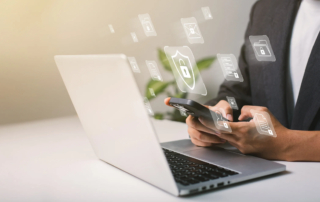 Responding to DarkSide ransomware threat. The image shows a person holding a smartphone while sitting in front of an open laptop. There are digital icons floating around the person, symbolizing various elements related to cybersecurity. These icons include a shield with a padlock in the center, folder icons with locks, and other data security symbols, all of which represent the secure handling of information. The person is dressed in business attire, with only their hands and part of their torso visible. The overall scene emphasizes the connection between mobile and computer security.