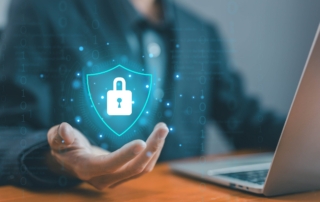 Business impact analysis for ransomware. This image shows a business professional in a suit sitting at a desk with a laptop. Hovering over their open hand is a glowing digital shield with a padlock icon in the center, symbolizing data protection and cybersecurity. The surrounding graphics include binary code and digital elements, reinforcing the theme of technology and security. The shield represents safeguarding sensitive information, and the setting suggests that this person is engaged in managing or enhancing cybersecurity measures in a professional, tech-driven environment.