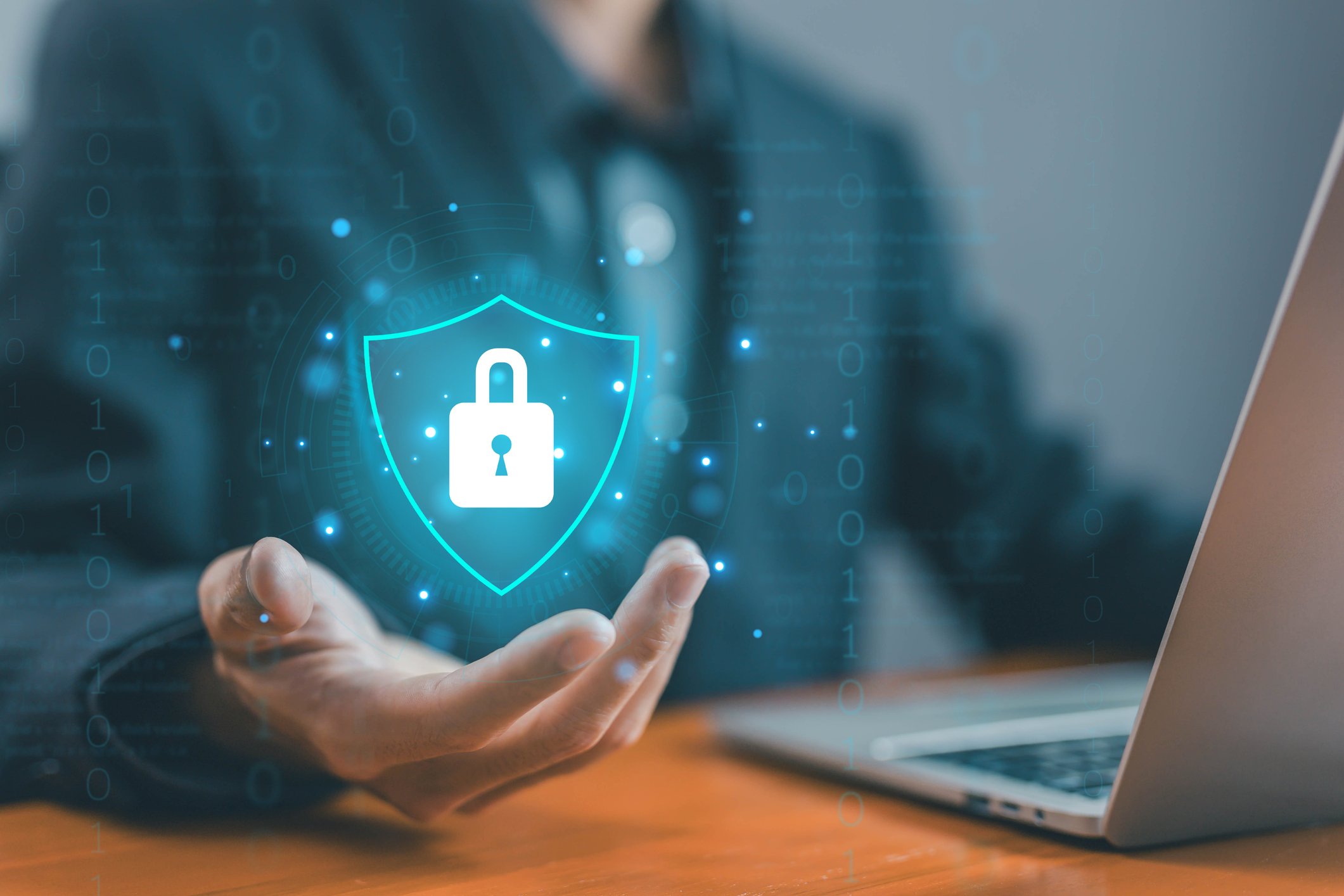 Business impact analysis for ransomware. This image shows a business professional in a suit sitting at a desk with a laptop. Hovering over their open hand is a glowing digital shield with a padlock icon in the center, symbolizing data protection and cybersecurity. The surrounding graphics include binary code and digital elements, reinforcing the theme of technology and security. The shield represents safeguarding sensitive information, and the setting suggests that this person is engaged in managing or enhancing cybersecurity measures in a professional, tech-driven environment.