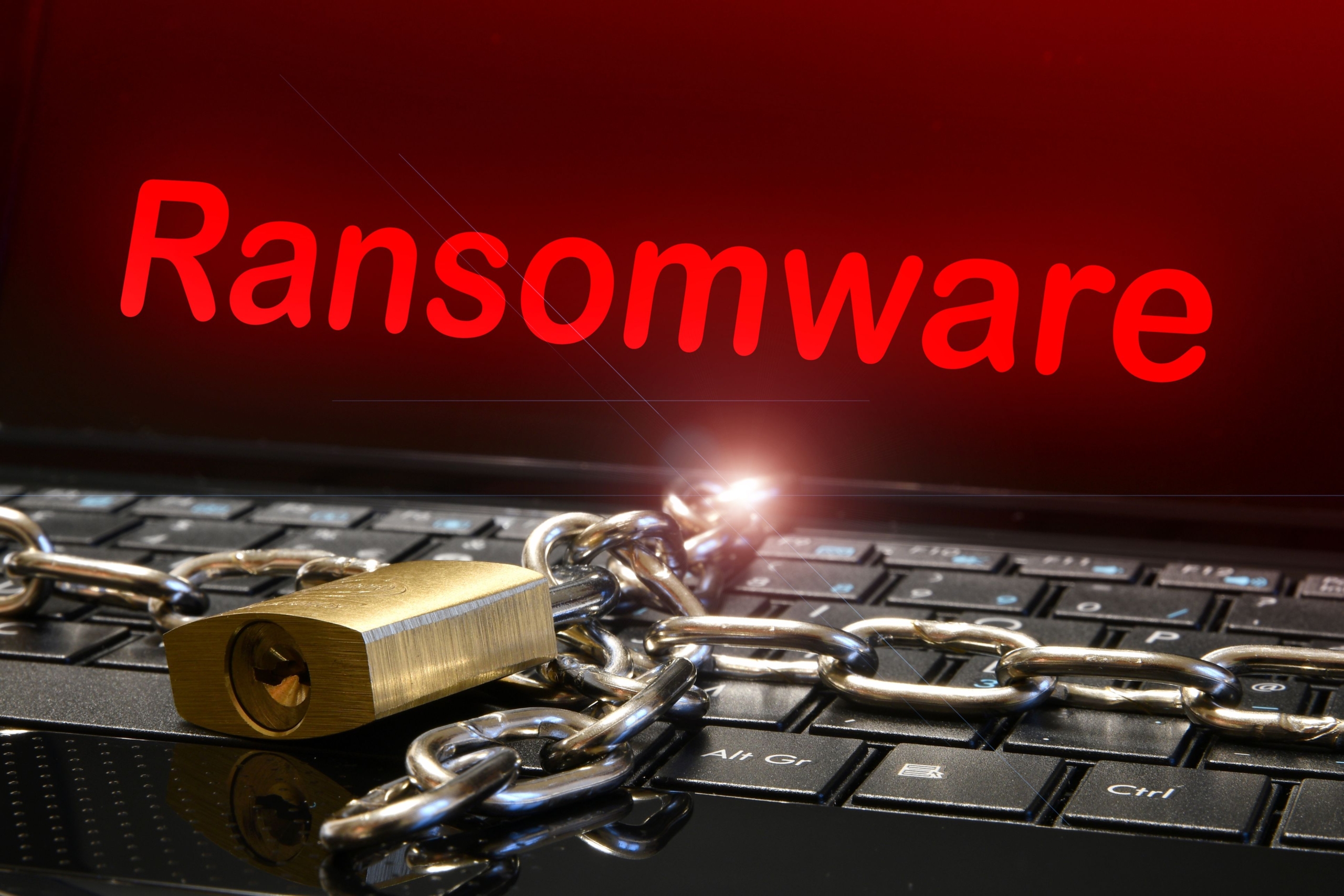 Business continuity plan for ransomware. The image visually represents a ransomware attack. The background shows a laptop keyboard, symbolizing the device being targeted. A heavy metal chain is draped across the keyboard, and a brass padlock secures the chain, symbolizing the device or data being locked or inaccessible. Above the keyboard, in bold, glowing red text, the word "Ransomware" is prominently displayed on a screen, creating a sense of urgency and threat. The red color emphasizes danger, while the chain and lock illustrate the concept of restricted access or encryption often associated with ransomware attacks. The overall tone is one of caution and cyber vulnerability.