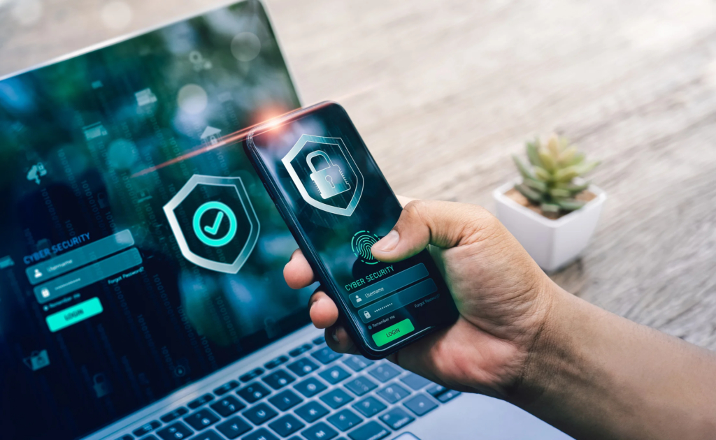 Cybersecurity resilience planning. The image shows a person holding a smartphone displaying a digital shield icon with a check mark, indicating security verification. The phone is positioned in front of a laptop, which also shows similar cybersecurity elements, including a login screen with fields for username and password. Both devices emphasize cybersecurity themes, with icons symbolizing authentication and data protection. The background includes a blurred workspace and a small plant, giving a professional and secure feel to the setup, likely representing two-factor authentication or secure login practices.