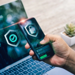 Cybersecurity resilience planning. The image shows a person holding a smartphone displaying a digital shield icon with a check mark, indicating security verification. The phone is positioned in front of a laptop, which also shows similar cybersecurity elements, including a login screen with fields for username and password. Both devices emphasize cybersecurity themes, with icons symbolizing authentication and data protection. The background includes a blurred workspace and a small plant, giving a professional and secure feel to the setup, likely representing two-factor authentication or secure login practices.