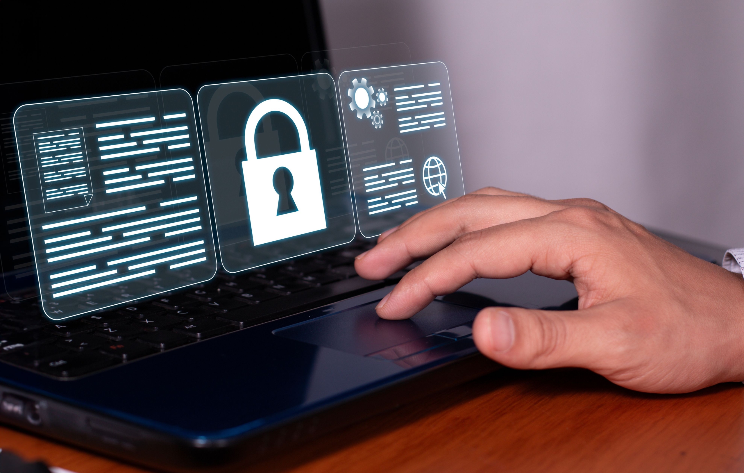 Preparing business for ransomware attack. The image shows a person's hand resting on the touchpad of a laptop, symbolizing interaction with technology. Floating above the laptop screen are three glowing, semi-transparent icons that resemble digital interface elements. The center icon features a large white lock, symbolizing security or protection of data. The icons on either side of the lock display digital text and symbols, resembling documents, encrypted files, or technical data. The overall design conveys themes of cybersecurity, data access control, and secure technology usage. The clean and futuristic look suggests a focus on digital solutions and online security measures. The neutral background keeps attention on the hand, laptop, and digital interface.