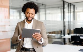 Ransomware attack communication plan. The image shows a well-dressed man standing in a modern office environment. He is smiling and holding a tablet, which he appears to be reading or interacting with. The man is wearing a light brown suit jacket over a white shirt, with a dark belt visible at his waist. His hair is curly, and he has a short beard. The background features glass walls and office furniture, giving the space an open and professional atmosphere. The overall tone of the image suggests a confident and engaged professional in a workplace setting.