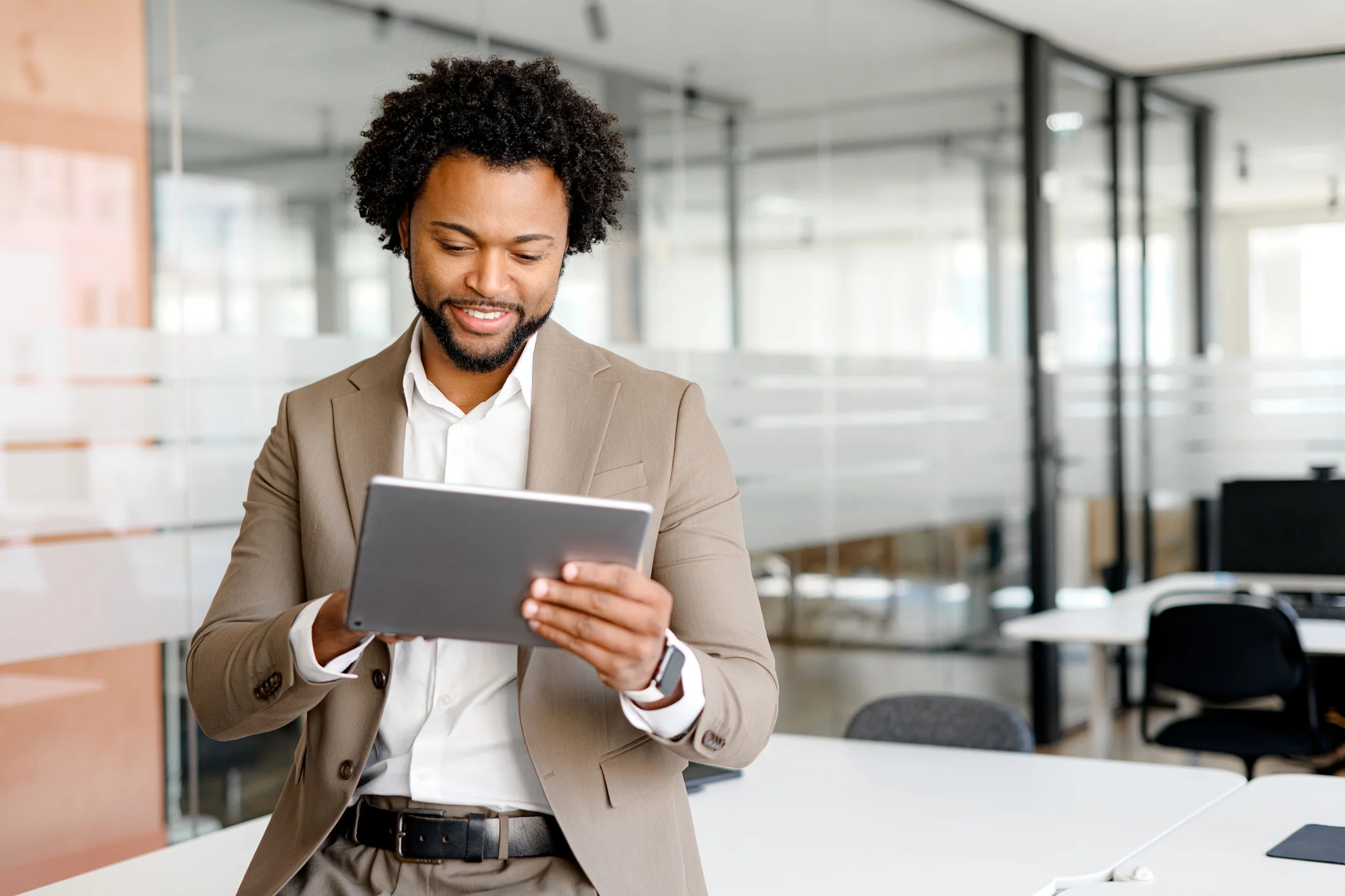 Ransomware attack communication plan. The image shows a well-dressed man standing in a modern office environment. He is smiling and holding a tablet, which he appears to be reading or interacting with. The man is wearing a light brown suit jacket over a white shirt, with a dark belt visible at his waist. His hair is curly, and he has a short beard. The background features glass walls and office furniture, giving the space an open and professional atmosphere. The overall tone of the image suggests a confident and engaged professional in a workplace setting.