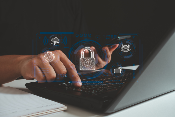 Business continuity planning for ransomware. This image features a person interacting with a laptop, their hand poised over the keyboard while pointing at the screen. Superimposed over the scene are digital, holographic-style graphics related to cybersecurity and data technology. The most prominent symbol is a glowing padlock in the center, symbolizing security. Surrounding it are icons representing a cloud, a fingerprint (likely denoting biometric access), a folder, and a database stack. The layout has a high-tech, futuristic vibe, emphasizing themes of secure data, digital storage, and technological advancement. The person is likely engaging in an activity related to these concepts, such as secure login or data management.