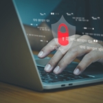 Handling REvil ransomware attack. This image shows a close-up of a person’s hands typing on a laptop keyboard. Overlaid on the image is a graphic representation of cybersecurity, featuring a red padlock icon set against a transparent shield shape. The padlock symbolizes security, and binary code (sequences of ones and zeros) is displayed floating in the background, further emphasizing the concept of digital protection. The overall tone of the image is serious and professional, with a dark, muted color scheme to evoke themes of technology and cybersecurity.