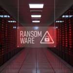 Recovering from Netwalker ransomware attack. This image depicts a data center with rows of server racks illuminated by red lights, creating an intense and alarming atmosphere. Hanging in the center of the image is a translucent sign displaying the word "RANSOMWARE" in bold white letters, accompanied by a warning triangle icon with a padlock inside it. The perspective draws attention to the central aisle, which is well-lit and contrasts with the red glow of the servers, emphasizing the theme of cybersecurity threats and the urgency of ransomware attacks. The overall mood is stark and ominous, fitting for the subject of digital vulnerabilities.