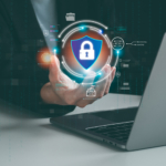 Encryption strategies against ransomware. A close-up of a hand reaching toward a floating holographic shield with a padlock at the center, displayed above a laptop. The background includes glowing digital data and network symbols, emphasizing online security and encryption.