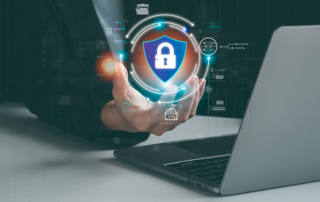 Encryption strategies against ransomware. A close-up of a hand reaching toward a floating holographic shield with a padlock at the center, displayed above a laptop. The background includes glowing digital data and network symbols, emphasizing online security and encryption.