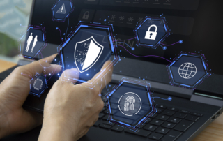 IoT device security from ransomware attacks. A pair of hands hovers over a laptop keyboard, with holographic icons floating above the screen. These icons include a shield symbolizing cybersecurity, a fingerprint for biometric authentication, a padlock for security, and a globe representing internet connectivity.