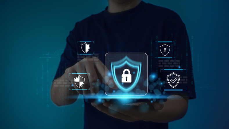 Third-party risk management for ransomware. A person sitting at a desk using a laptop. Their hand points towards a glowing shield icon with a padlock at its center. A web of connected icons surrounds the shield, representing cybersecurity, digital networks, and data protection.