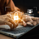 Cybersecurity war room strategies. A person is typing on a laptop keyboard, wearing an earth-toned sweater. A golden, glowing security shield icon with a padlock is digitally overlaid on the hands. Additional icons appear, including a user login interface, symbolizing online security or access control. The background is dark and slightly blurred, with soft lighting highlighting the hands and laptop. Small floating, glowing security icons add a tech-heavy cybersecurity theme.