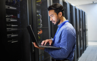 Economics of ransomware attacks. A man with short black hair and a beard, wearing a blue button-up shirt and an ID badge, is working on a laptop in a data center. He is standing next to tall server racks filled with network cables and blinking lights, suggesting an IT professional or cybersecurity specialist. The environment is modern, highly secure, and technology-driven.