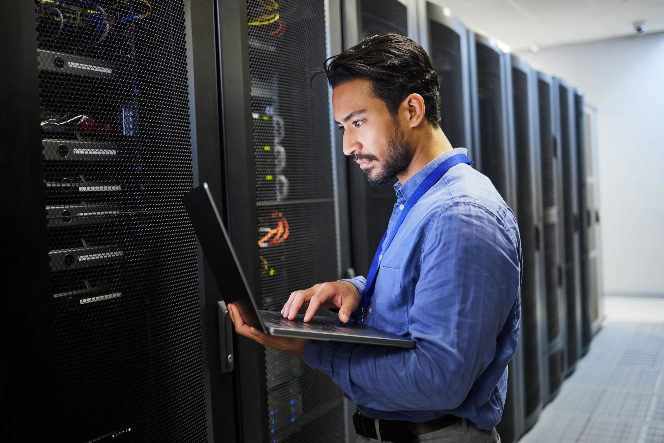 Economics of ransomware attacks. A man with short black hair and a beard, wearing a blue button-up shirt and an ID badge, is working on a laptop in a data center. He is standing next to tall server racks filled with network cables and blinking lights, suggesting an IT professional or cybersecurity specialist. The environment is modern, highly secure, and technology-driven.