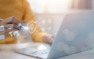 Ransomware protection for financial institutions. A person wearing a mustard-colored sweater is using a stylus on a laptop. A translucent, glowing digital interface appears, featuring a shield with a keyhole, a world map, financial icons, and a database, suggesting a focus on cybersecurity, financial security, and global data management.