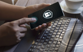 Ransomware-resistant file systems. A person is holding a smartphone horizontally with both hands. The phone’s screen displays a padlock symbol inside a shield, indicating security or encryption. The hands appear well-lit and natural, with fingers touching the screen. Below the hands is a black keyboard on a reflective surface, likely a laptop. In the background, there’s a white coffee cup on a saucer, suggesting a casual work or café setting. There are subtle digital effects, such as faint circles and glowing edges around the padlock.