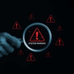 Data loss prevention in ransomware defense. A hand holding a magnifying glass is examining a screen filled with red exclamation marks inside warning triangles. The text "System Warning" is displayed in the center, indicating a critical error, vulnerability, or cybersecurity alert. The background is black with a hacker-style digital threat environment, emphasizing cybersecurity risks.