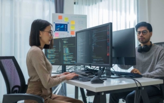 Backup solutions ransomware resilience. Two young professionals sit across from each other at a shared desk in a bright, minimalist office space. Each person has multiple computer monitors displaying lines of code. A woman on the left is typing, while a man on the right, wearing glasses and headphones around his neck, looks attentively at his screen. Colorful sticky notes are visible on a board in the background.