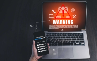 Cloud ransomware vulnerability assessment. A hand holds a smartphone above an open laptop. On the laptop screen is a vivid red warning symbol with an exclamation mark inside a triangle, along with icons for email and settings. White arrows labeled "IN" and "OUT" point between the phone and laptop, suggesting a file upload or data transfer in progress. The scene conveys a cyberattack, security breach, or phishing warning.