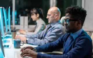 Cybersecurity Awareness Training. Three people sit side-by-side at a long desk, each working on desktop computers. They appear focused and professional. A man with glasses and curly hair in the foreground types attentively. Behind him, a bald man and a woman with her hair tied back are also concentrating on their screens. The setting appears to be a modern office or cybersecurity operations center, with a cool blue lighting.