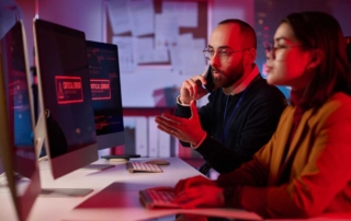 Employee Cybersecurity Training Programs. Two cybersecurity professionals—a man and a woman—sit at their desks in a red-lit room. They are discussing something on their screens, which show alerts labeled “CRITICAL ERROR.” The man is speaking on the phone while gesturing toward the screen.