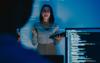Proactive Infrastructure Monitoring. A woman stands in a dark room, illuminated by a blue glow from projected code or data on the wall behind her. She holds a tablet and appears to be giving a technical presentation. Part of a computer screen showing lines of code is visible in the foreground, slightly blurred.
