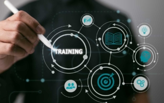 Real-Time Network Monitoring. A close-up of a hand holding a stylus or pen, pointing at a futuristic digital interface. The word "TRAINING" is highlighted in the center of interconnected icons: a target, a gear, a lightbulb, an open book, and groups of people. The visual suggests concepts of learning, skill development, and structured knowledge building in a digital or corporate environment.