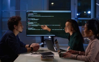 Secure Data Backup Solutions. Three colleagues sit around a table in a dimly lit office with large windows behind them showing nighttime city lights. They are engaged in a discussion in front of a large computer monitor displaying code. One woman is pointing at the screen while speaking, while another woman and a man follow along attentively with laptops and smartphones in hand. There are notebooks and glasses on the table.