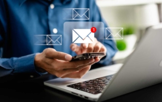 VPN ransomware protection strategies. A close-up image shows a person in a blue button-down shirt using a smartphone while sitting at a desk with a laptop open. Floating above the phone are transparent email icons, and one email has a red notification badge, symbolizing a new incoming message. The setting appears modern and professional.