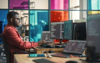 Automated Software Update Services. A man sits in a colorful tech office, focused on coding across multiple monitors in a modern, vibrant workspace.