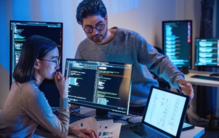 Data Protection Backup Strategies. Two people collaborate in a dark-lit room full of screens with code; one points to a tablet while the other reviews documents, immersed in technical problem-solving.