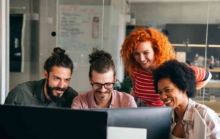 Email Threat Detection Solutions. Four colleagues huddle around a computer, laughing and smiling together, capturing a moment of friendly teamwork in an open office.