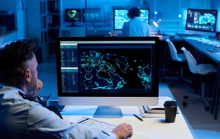 Patch Management for Compliance. A man sits at a desk in a dimly lit control room, facing a large monitor displaying a global cyber map with glowing data points. Multiple screens line the room, and another person works in the background. The environment feels tense and analytical, like a command center monitoring cyber threats in real time.