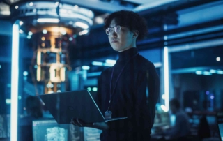 Rapid Disaster Recovery Solutions. A young person with glasses stands confidently in a high-tech data center, holding a laptop. Bright blue and white LED lights illuminate the room, casting a futuristic glow. It’s a scene of high-level tech work in a secure environment.