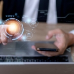 byod-endpoint-protection-strategies BYOD Endpoint Protection Strategies. A businessperson pointing at a glowing orange fingerprint scanner hologram while holding a smartphone, with faint futuristic interface graphics around the image.