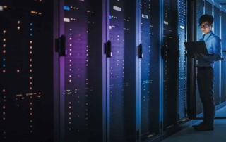 Cybersecurity Mesh Solutions. A person stands in a server room lined with glowing, tall server racks, holding a laptop and checking the equipment.