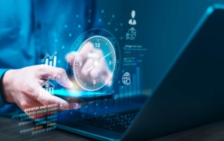 Enterprise Vulnerability Management. A hand interacts with a tablet in front of a laptop. The screen emits a glowing digital clock and data charts. It suggests time-sensitive data processing, real-time monitoring, or system uptime.